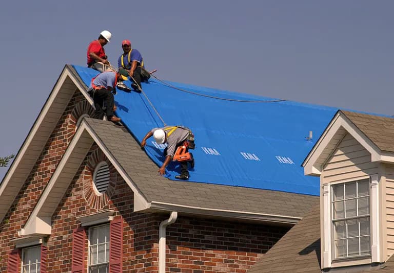 Four crew members securing a FEMA-marked blue tarp on a brick home's roof