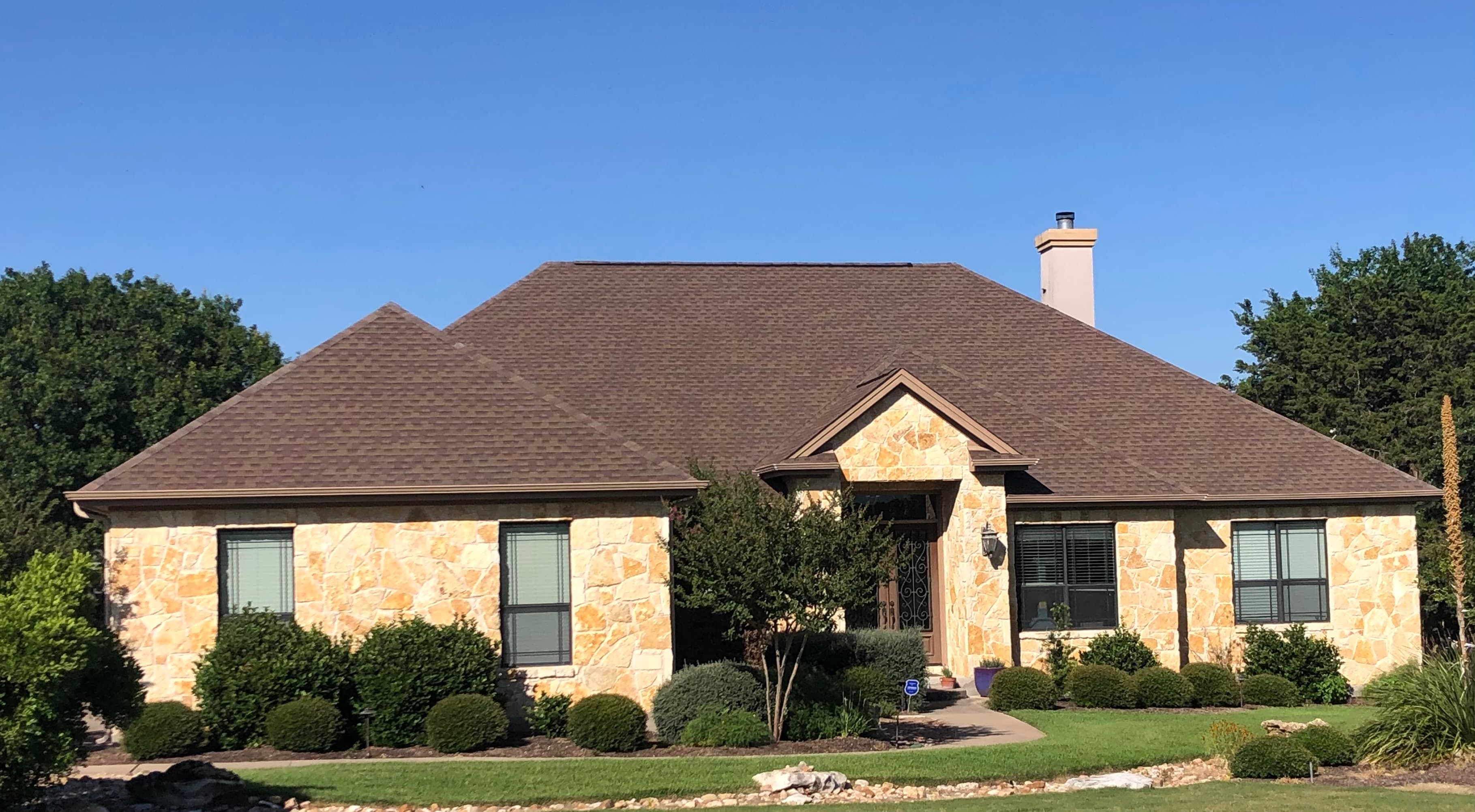 Stone home with a bronze-tone architectural shingle roof under a clear blue sky